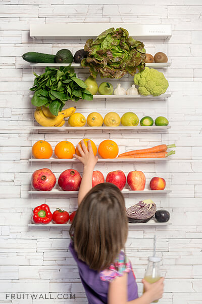 Frutero de pared colgante con 6 estantes lleno de frutas y verduras ordenadas por colores en forma de arcoiris con una niña que coge una naranja mientras bebe un zumo verde