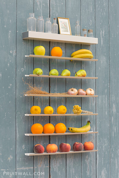 Frutero de pared colgante con 6 estantes sobre fondo de madera gris lleno de naranjas, manzanas, melocotones, peras, limones, cebollas, platanos, flores y botes de cristal